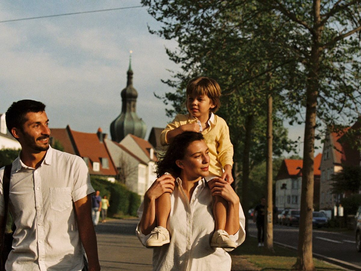 Eine Familie geht am späten Nachmittag eine Allee in einer Kleinstadt entlang, das Kind sitzt auf den Schultern
