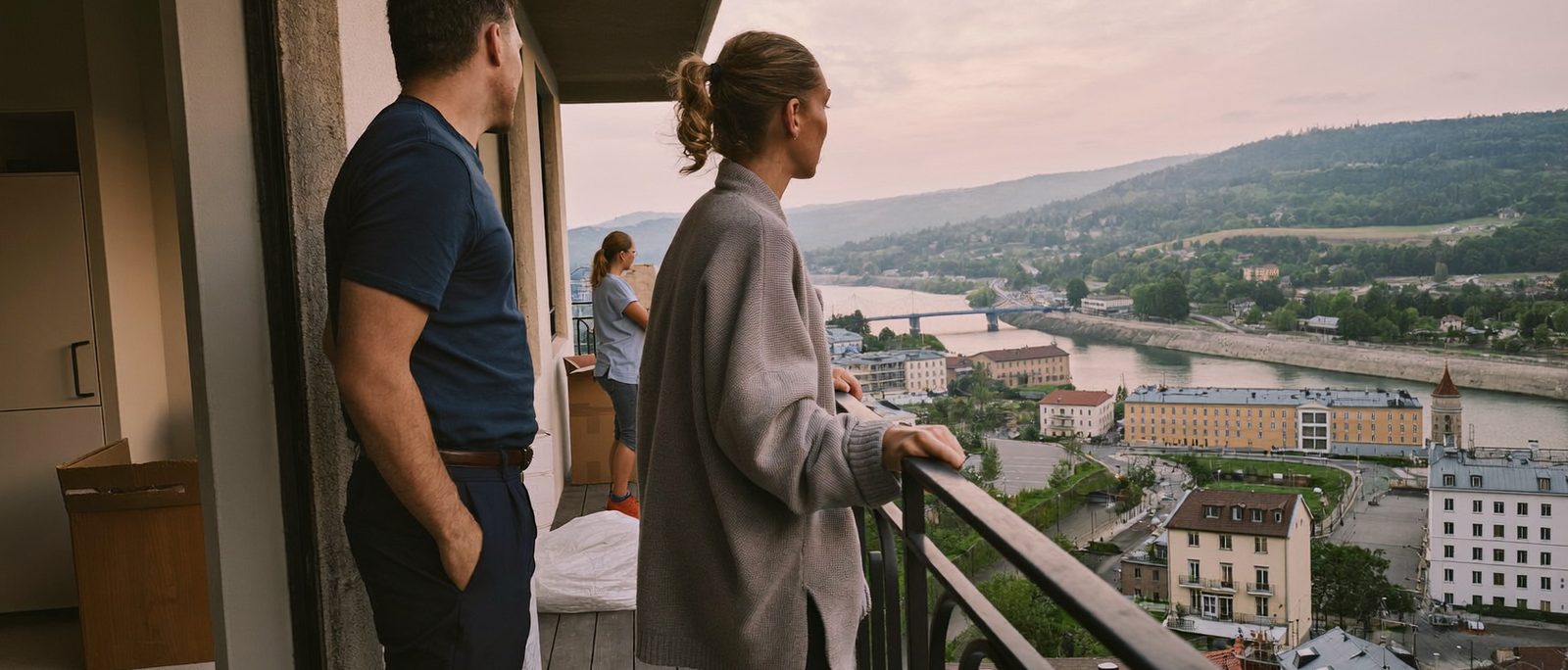 Eine Familie steht am Abend auf dem Balkon einer neuen Wohnung und blickt über eine Stadt am Fluss, Umzugskartons im Hintergrund